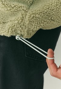 Roheline koheline fliisriie tekstuurse pinnaga, millel on metallist klõps ja valge tõmbelint musta taustal.