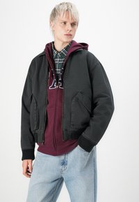 Jongere in lichtblauwe jeans, maroon hooded jacket met letters, groen geruit overhemd en zwarte jas, staande met één hand in de zak.