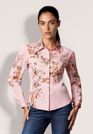 Frau mit dunklem zurückgebundenem Haar, die ein enganliegendes rosa geblümtes Hemd mit Knöpfen und blaue Jeans trägt, posiert vor einem schlichten hellen Hintergrund.