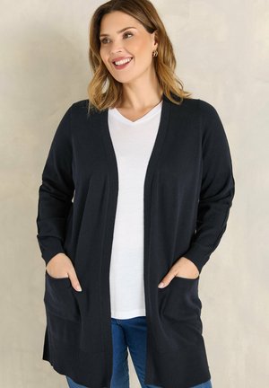 Donna sorridente che indossa un cardigan nero aperto sopra una maglietta bianca con scollo a V e jeans blu, con le mani nelle tasche del cardigan, in piedi davanti a uno sfondo neutro.