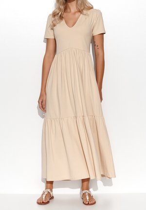 Vrouw draagt een beige maxi-jurk met korte mouwen en V-hals, met geplooide rokpanelen en witte sandalen met strik, staand tegen een witte achtergrond.