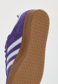 Sapatilha de camurça roxa com riscas brancas e colarinho acolchoado. O solado em borracha apresenta um padrão hexagonal. Perfil lateral com detalhes de costura notáveis.