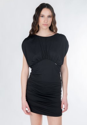 Jonge vrouw met lang golvend haar, gekleed in een aansluitende zwarte mini-jurk met plooien, staand tegen een effen witte achtergrond.