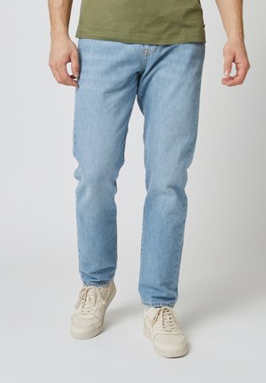 Man die een lichtblauwe spijkerbroek, een olijfgroen T-shirt en gebroken witte sneakers draagt, staat tegen een egale lichte achtergrond.