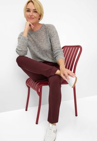 Grau strukturierter Pullover, burgunderfarbene Hose und weiße Sneakers. Modell sitzt auf einem roten Lattenstuhl mit neutralem Hintergrund.