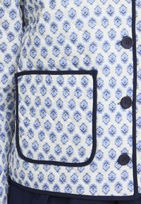 Secțiune de jachetă matlasată cu model floral albastru, margini și buzunar cu bandă albastru închis și trei nasturi albastru închis pe fundal alb.