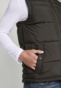 Dunkelgrüne Steppweste mit gestepptem Muster, Reißverschluss vorne und Druckknopf-Seitentaschen. Über einem weißen Langarmshirt getragen.