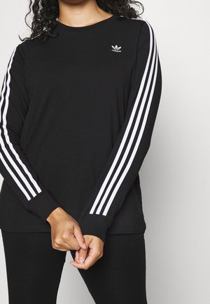 Personne portant un t-shirt noir à manches longues Adidas avec trois bandes blanches sur chaque manche et le logo Adidas sur la poitrine.