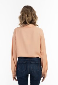 Langærmet ferskenfarvet bluse med krave, let glans og knaplukning ved håndleddene, parret med mørkeblå tætsiddende jeans med baglommer.