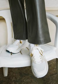 Weisse Sportschuhe mit Netz- und Lederelementen, goldenen Akzenten und weissen Schnürsenkeln, auf einem Plastikstuhl platziert, zusammen mit grünen Hosen.
