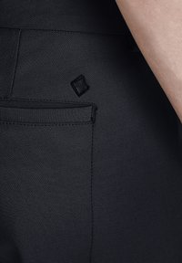 Zwarte op maat gemaakte broeken met een gladde structuur, voorzien van een achterzak en een klein zwart logo patch boven de zak.