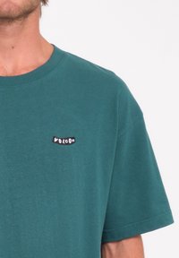 Homme portant un t-shirt Volcom teal, visible du cou au milieu du torse, fond blanc uni.