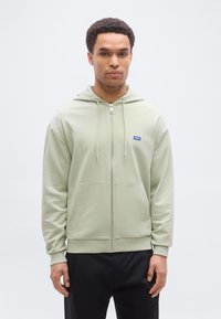 Νεαρός άνδρας που φοράει ανοιχτοπράσινο φούτερ με φερμουάρ και μπροστινές τσέπες, καθώς και μαύρο παντελόνι, στέκεται μπροστά σε ένα απλό λευκό φόντο.