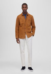 Chaqueta de ante marrón con cierre de botón y bolsillo en el pecho, llevada sobre una camisa azul, combinada con pantalones color crema y zapatillas negras.
