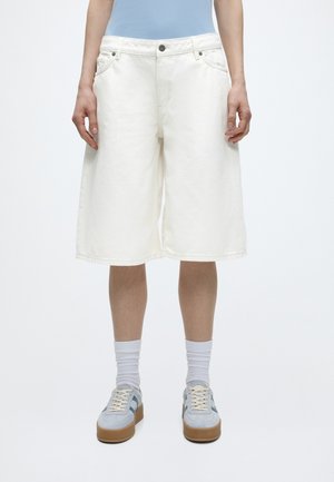 Alsó test laza, fehér térdig érő rövidnadrágot, fehér zoknit és világoskék, gumitalpú sportcipőt visel, fehér háttéren áll.