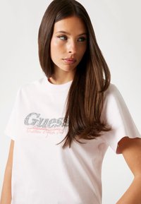 Tricou din bumbac roz, cu un logo argintiu strălucitor "Guess" și textul roșu "Nineteen Eighty One". Mâneci scurte și croială lejeră.