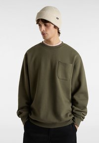 Olivengroene sweatshirt met een ronde hals, lange mouwen en een borstzak; gedragen met een lichtbeige geribde beanie.