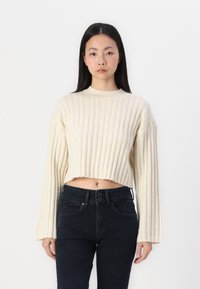 Kremfarget ribbestrikket cropped genser med lange ermer, med høy hals. Satt sammen med mørke høytaljede jeans.