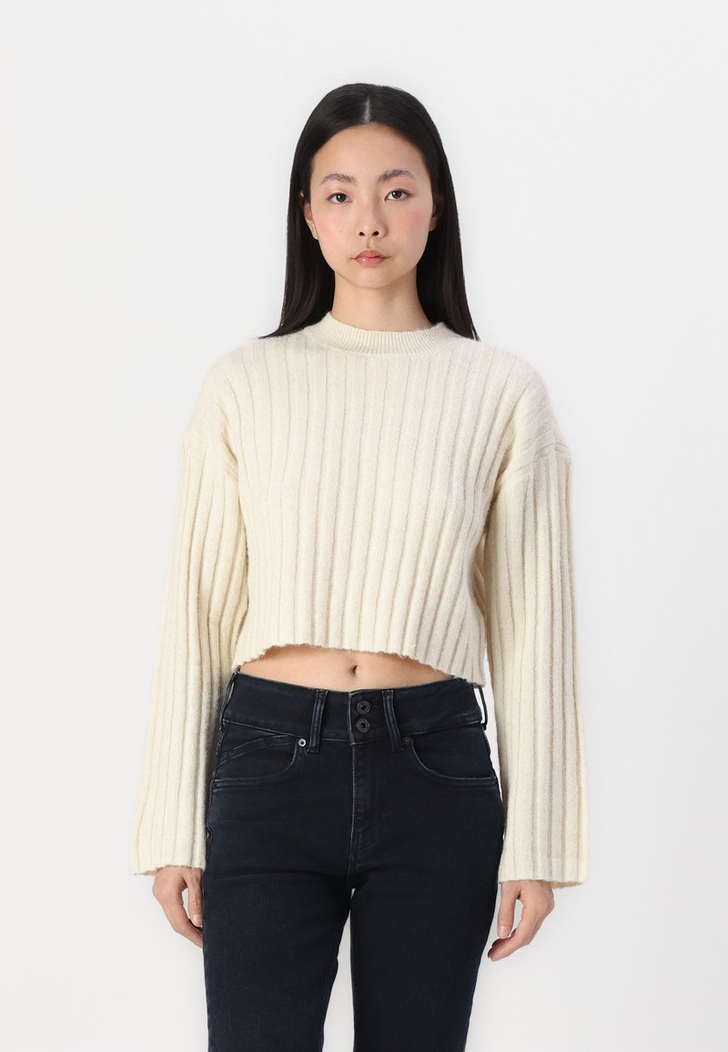 Kremfarget ribbestrikket cropped genser med lange ermer, med høy hals. Satt sammen med mørke høytaljede jeans.