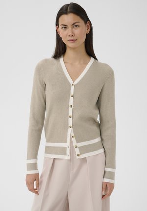 Naine, kes kannab beeži soonikkoes kardigani valgete ääriste ja nööpidega, kooshele valgelt beežide laia säärega pükstega, seisab ühevärvilise tausta ees.