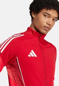 Rote Sportjacke mit hohem Kragen, weißem Drei-Streifen-Design an den Ärmeln, Reißverschluss vorne und strukturiertem Einsatz an der Seite.