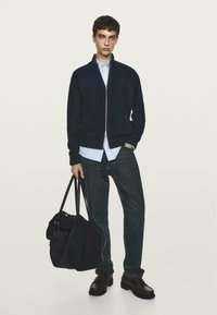 Maglione zip-up navy, camicia azzurro chiaro sotto, jeans scuri, borsa nera, scarpe marroni, modello in piedi contro uno sfondo leggero e uniforme.