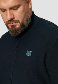 Marineblauer Strick-Reißverschluss-Pullover mit hohem Kragen, ausgestattet mit einem hellblauen gestickten Logo auf der linken Brust und strukturiertem Stoff.