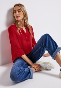 Blusa rossa a maniche lunghe con bottoni, abbinata a jeans in denim blu a taglio rilassato con risvolto e sneakers bianche, seduta contro un muro.