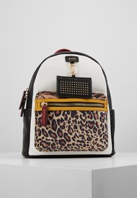 Mochila compacta con paneles en blanco, negro y estampado de leopardo, un bolsillo frontal con cremallera y borde mostaza, y una etiqueta de llavero negra con tachuelas.