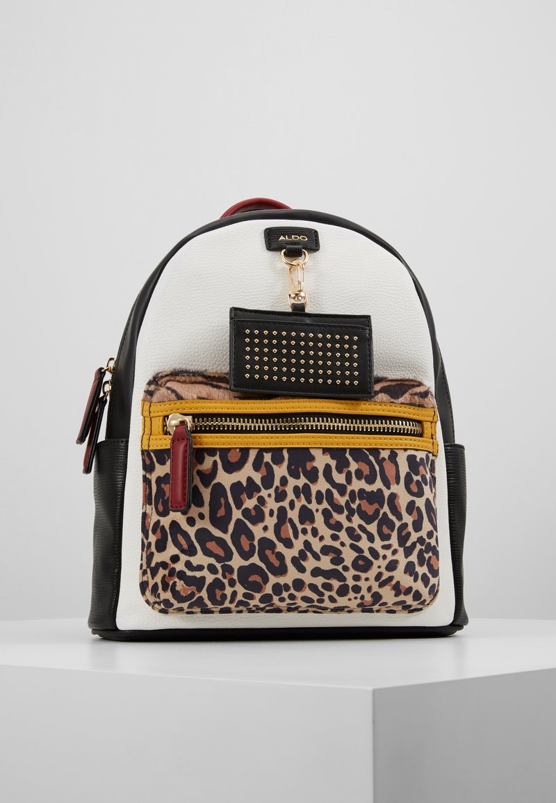 Mochila compacta con paneles en blanco, negro y estampado de leopardo, un bolsillo frontal con cremallera y borde mostaza, y una etiqueta de llavero negra con tachuelas.