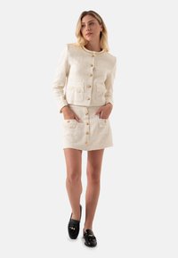 Weißes strukturiertes Jacket mit goldenen Knöpfen und passendem Rock, beide mit aufgesetzten Taschen. Schwarze Loafers runden das Outfit ab.