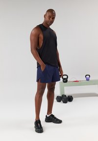 Black sleeveless athletic top, navy shorts with side slits, and black sports shoes. Background includes kettlebells and dumbbells.