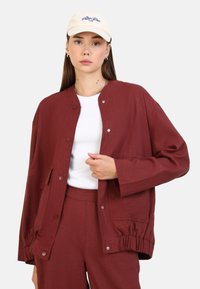 Veste bordeaux avec une coupe décontractée, dotée de boutons-pression, de poches latérales et de poignets froncés. Portée par-dessus un haut blanc et assortie à un pantalon.