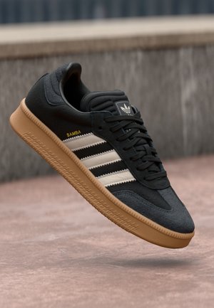 Sort Adidas Samba sneakers med hvide striber og gummisål, der svæver over en betonoverflade med en grå væg i baggrunden.
