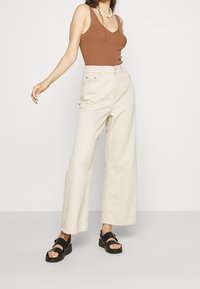 Femme portant un haut marron sans manches, un pantalon beige taille haute à jambes larges, des sandales noires et un collier blanc en perles, debout avec le bras tendu.