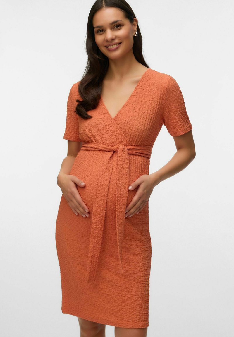 Femme enceinte souriante portant une robe portefeuille orange texturée, les mains posées doucement sur son ventre, avec de longs cheveux foncés tombant sur une épaule.