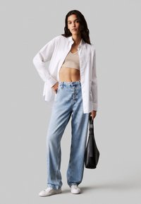 Camisa blanca de botones sobre un top corto beige, combinada con jeans de pierna ancha de lavado claro y zapatillas blancas. Sosteniendo un bolso negro.