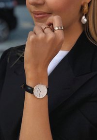 Round watch with a white dial, gold accents, and black leather strap. Accompanied by rings and gold earrings with pearly details.