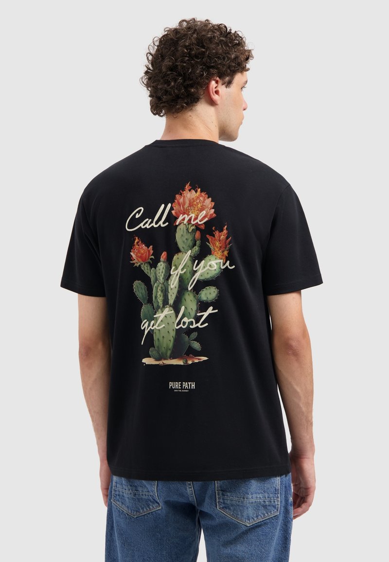 Jonge man met krullend haar die een zwart T-shirt draagt met een grote cactus met oranje bloemen en de tekst "Bel me als je verdwaald bent."