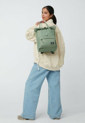 Young woman wearing beige jacket and wide blue jeans carries green roll-top backpack over one shoulder.