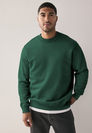 Felpa verde in cotone con collo rotondo, polsini e orlo a costine. Indossata su una camicia bianca, presenta una texture liscia e una vestibilità regolare.