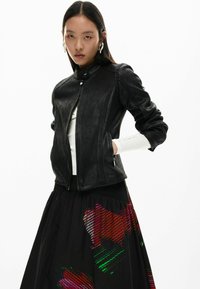 Must nahast jakk, millel on keha järgnev lõige, krõpsuga krae, küljetaskud ja sooniline tekstuur. Kantud valge topi ja mustrilise musta seelikuga.