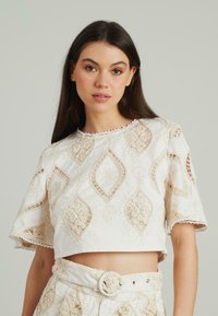 Witte crop top met ingewikkelde beige borduursels, met bloemen- en diamantpatronen, golvende halslijn en korte mouwen.