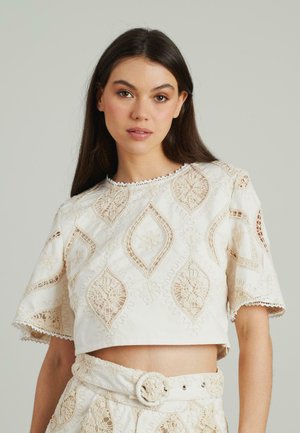 Top corto bianco con intricati ricami beige, caratterizzato da motivi floreali e a diamante, scollatura a scallop e maniche corte.