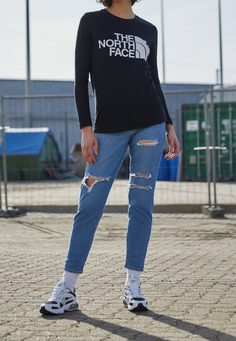 Chemise noire à manches longues avec le logo "The North Face", jean bleu clair déchiré, et baskets blanches avec des accents noirs et gris.