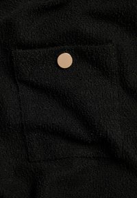 Svart texturerad tyg med en liten rektangulär ficka och en cirkulär knapp i guldton. Ytan har en mjuk, boucle-liknande känsla.