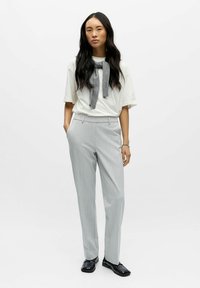 Kobieta z długimi ciemnymi włosami, ubrana w białą koszulkę, jasnoszare spodnie, czarne buty i szary sweter narzucony na ramiona.