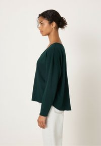 Tumši zaļš brīvs džemperis ar V veida kakla izgriezumu, garām piedurknēm un brīvu piegriezumu, izgatavots no mīkstas auduma, attēlots uz gaiša fona.