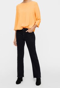 Orange, locker sitzende Bluse mit Dreiviertelärmeln, kombiniert mit schwarzen Schlagjeans. Modell trägt schwarze Knöchelstiefel. Einfaches Design, glatte Textur.