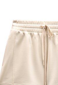 Beżowe spodenki z elastycznym pasem, bocznymi kieszeniami oraz wiązanym troczkiem z przodu, pokazane na białym tle.